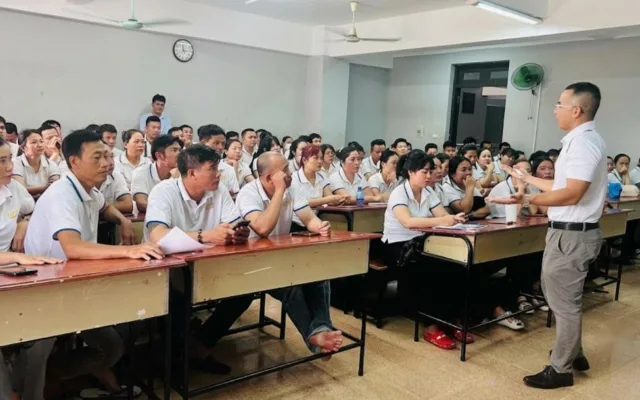 Người đứng giảng trước lớp đông người mặc áo polo trắng ngồi ở bàn học – cơ hội du học nghề Đức nhờ tiếng Đức