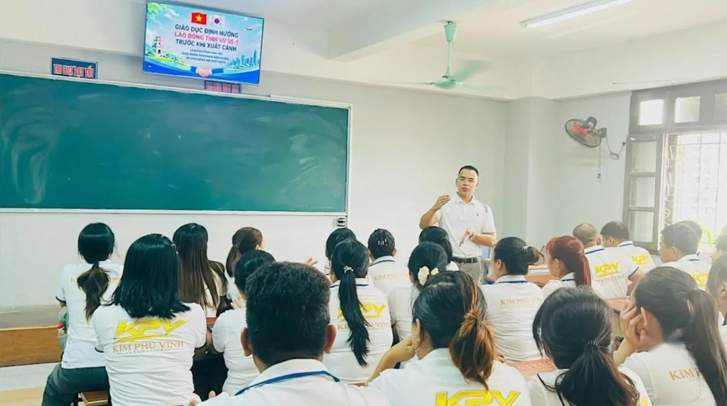 Người đứng giảng cạnh bảng xanh, học viên áo đồng phục trắng ngồi nghe trong lớp – không gian học tập, tư vấn tại KPV