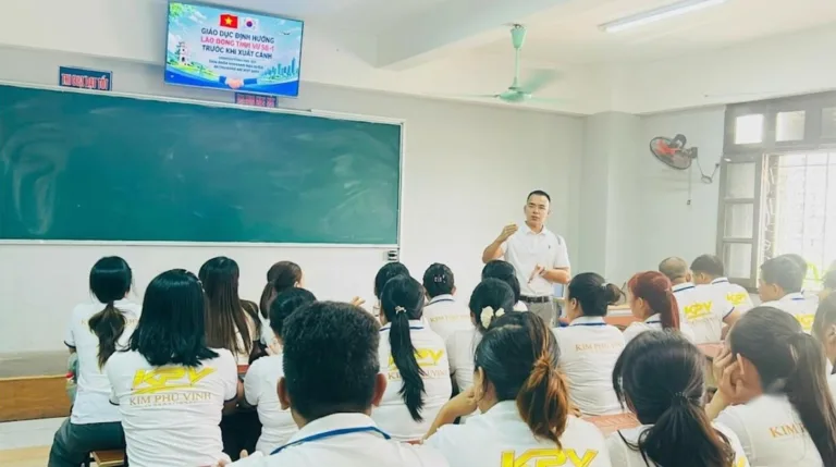 Người đứng giảng cạnh bảng xanh, học viên áo đồng phục trắng ngồi nghe trong lớp – đào tạo ngoại ngữ tại KPV Đồng Nai