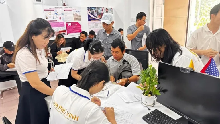 Nhân viên mặc áo KPV hướng dẫn mọi người điền hồ sơ tại bàn trong văn phòng, tường có poster chương trình du học