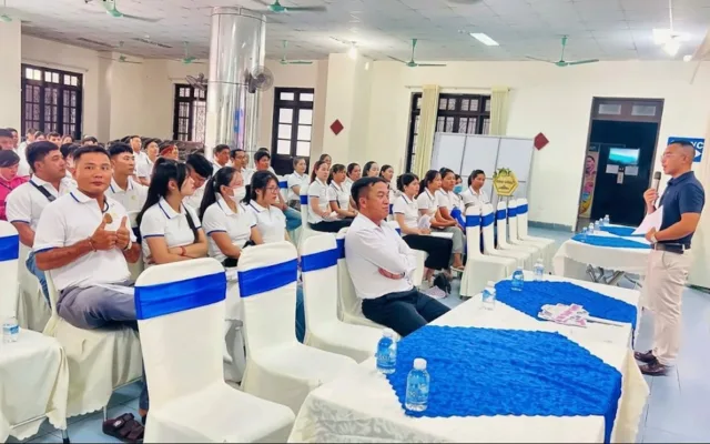 Người cầm micro phát biểu trước hội trường đông người ngồi nghe, ghế bọc trắng thắt nơ xanh – hội thảo định hướng KPV