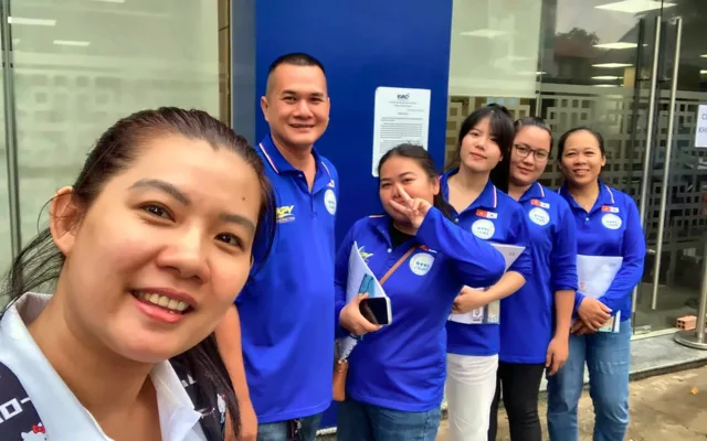 Nhóm sáu người áo xanh đồng phục chụp ảnh chung trước cửa kính KVAC – văn phòng tư vấn của KPV Group