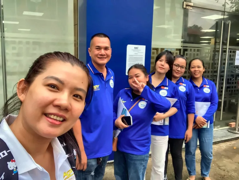 Nhóm sáu người áo xanh đồng phục chụp ảnh chung trước cửa kính KVAC – KPV Group (Kim Phú Vinh International)